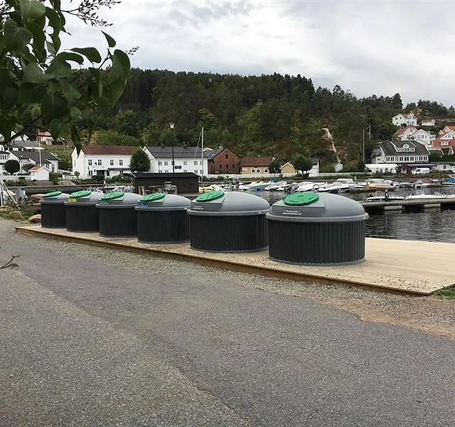 Rekke med SemiQ avfallsbeholdere ved båthavn, integrert i maritimt miljø med brygge og småhus i bakgrunnen.