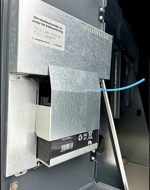 Metallbeskyttet batteriboks installert inne i avfallscontainer, med kabel og produsentens etikett synlig.