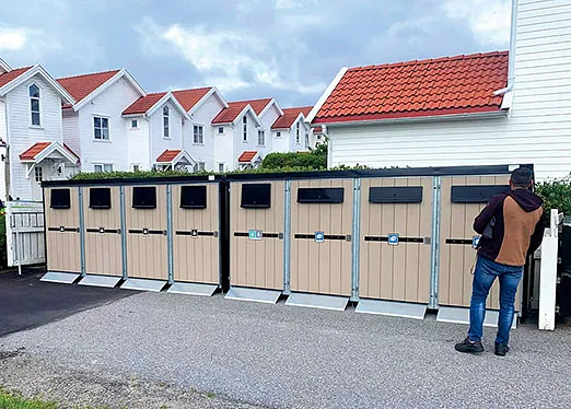 Flere beige skapmoduler tilpasset plastbeholdere, plassert i et boligområde med hvite hus og røde tak, en person står ved siden av.