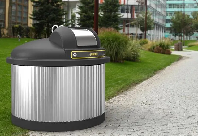 Delvis nedgravd SemiQ avfallsbeholder for plast, plassert i urbant grøntområde med moderne bygg i bakgrunnen.