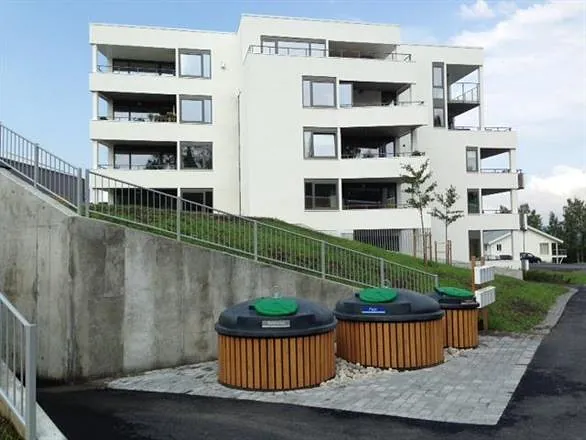 Tre SemiQ beholdere med trekledde paneler plassert ved moderne borettslag med hvit fasade og støttemur.