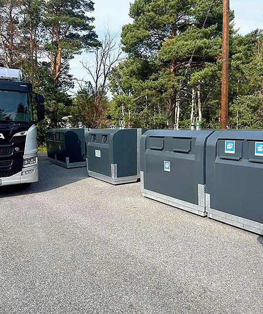 Overbakken containere av typen OWS Scandinavia i rekke med ulike sorteringsåpninger, plassert på asfalt foran lastebil og gjerde.